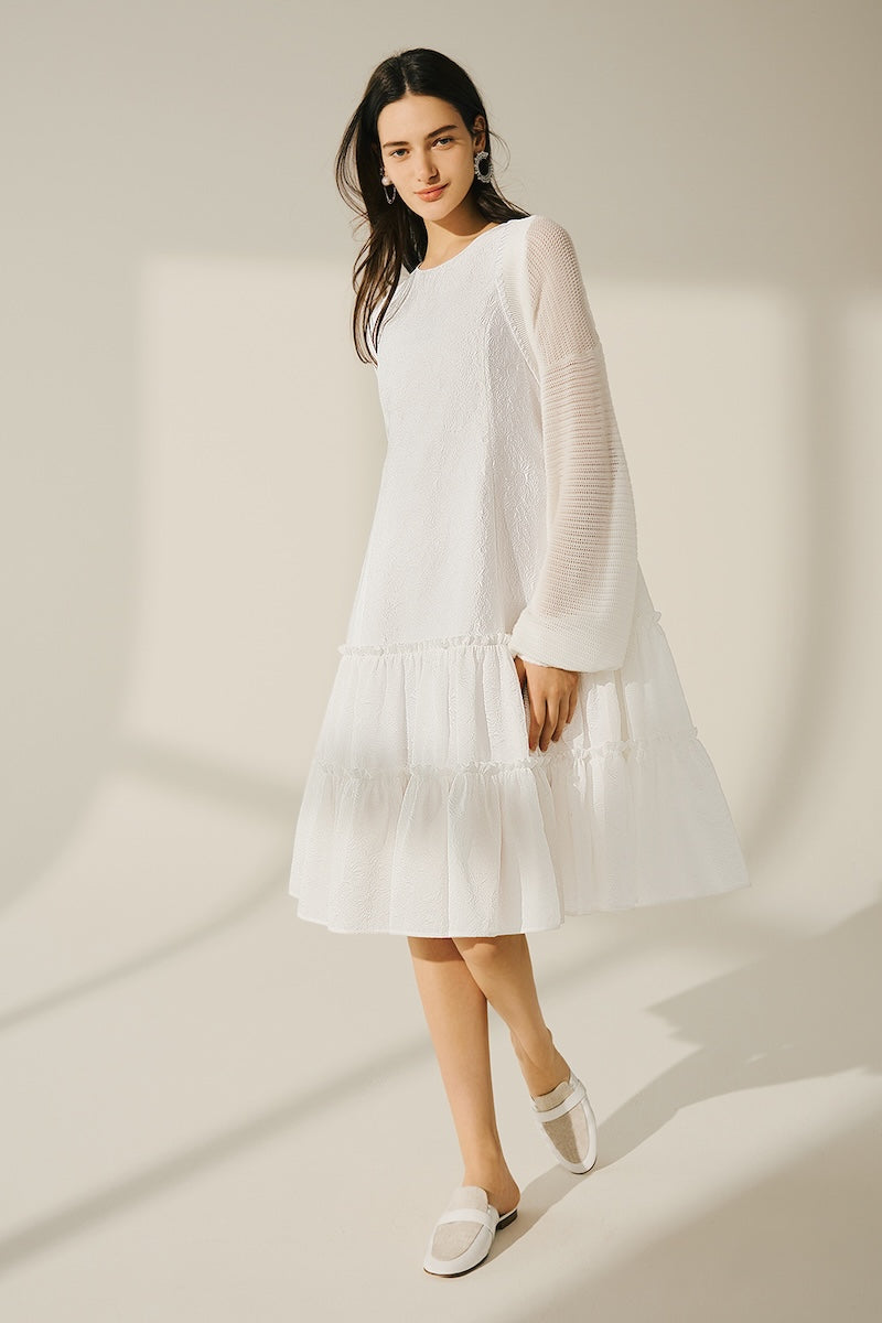 Model wearing a loose white long-sleeve dress with a tiered skirt and white flat shoes, standing in a softly lit room.