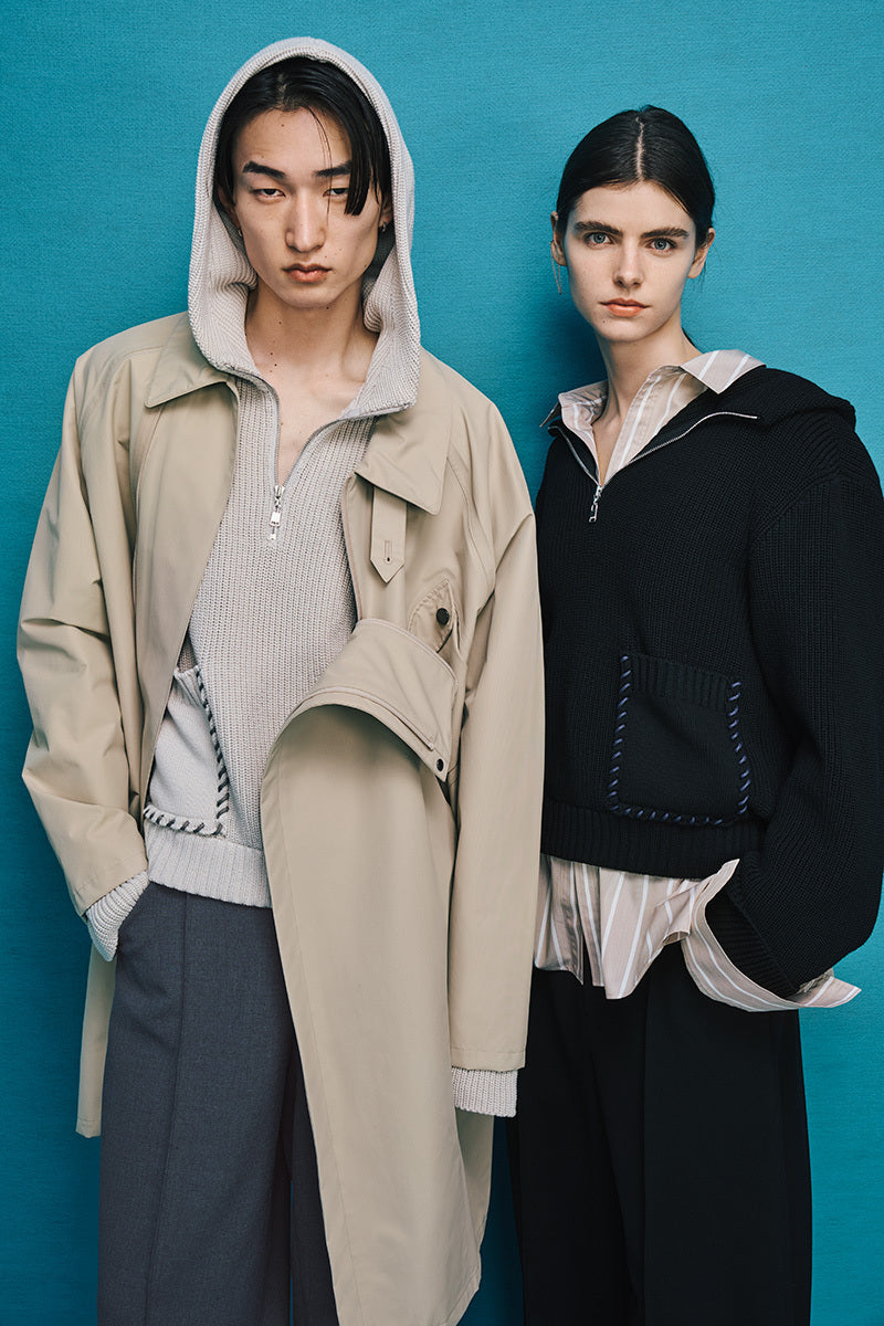 Two models are standing side by side. Model on the left is wearing dark gray pants with a beige coat over a beige top with a hood. Model on the right is wearing black pants with a black jacket over a beige shirt.