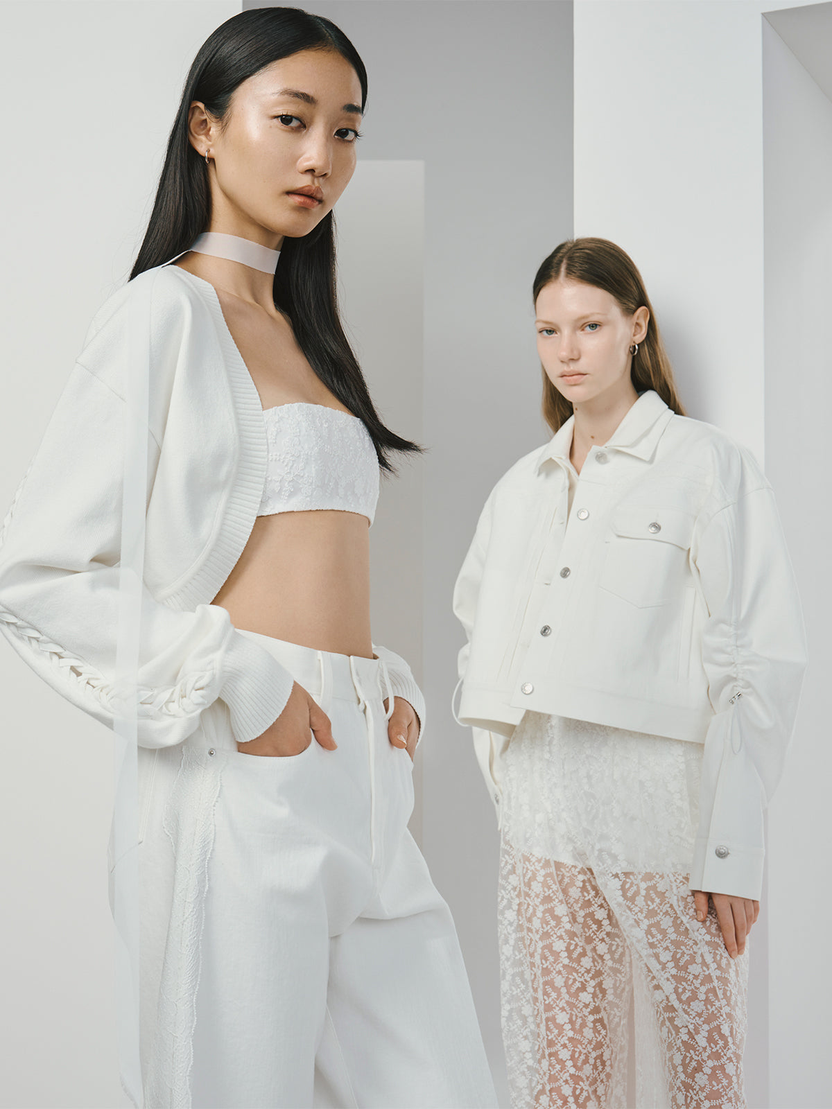 Two women wearing modern all-white outfits; one in a cropped bandeau top with wide-leg pants and a cardigan, the other in a cropped jacket over a sheer lace dress, standing against a minimal white background.