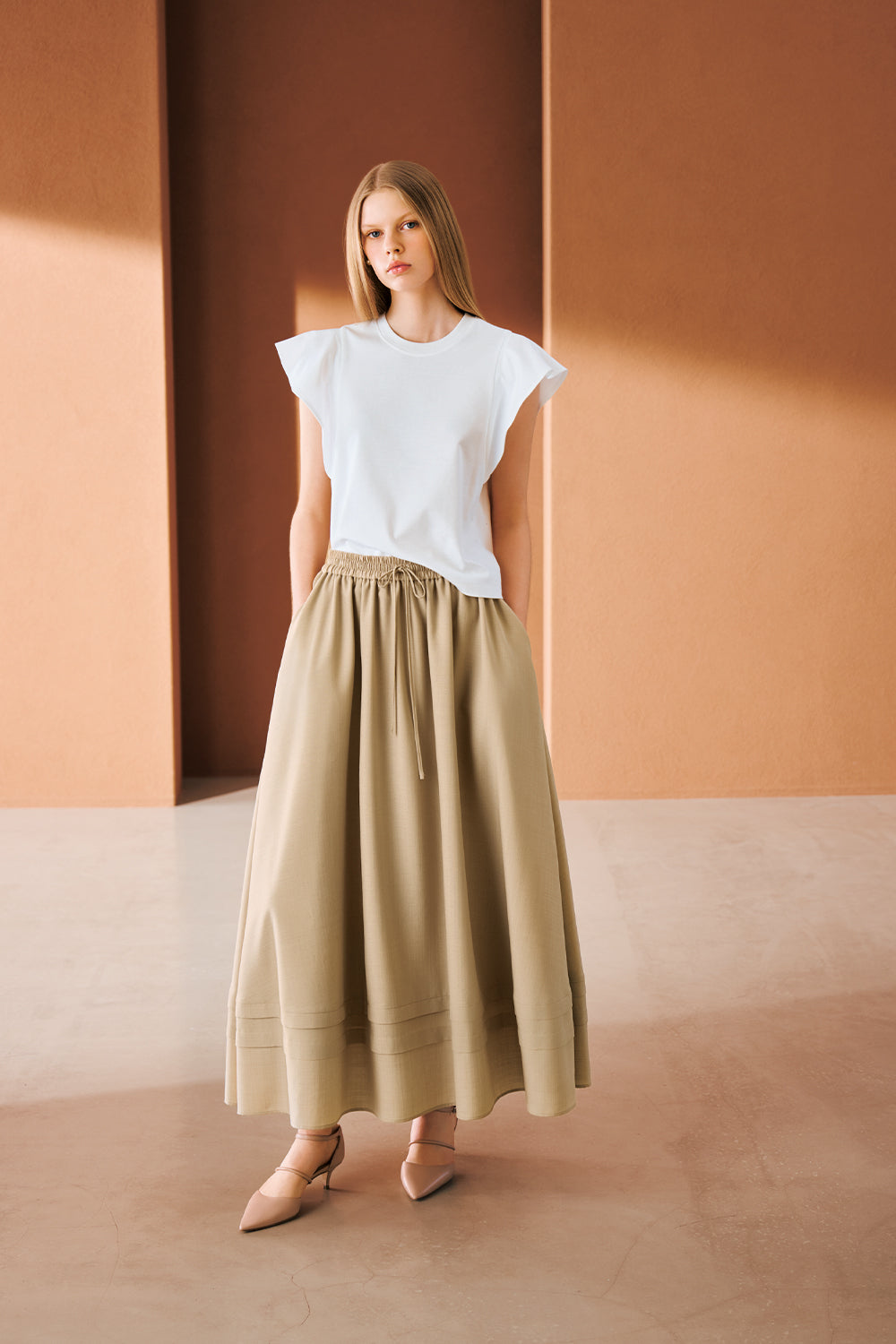 Model in a white crew-neck T-shirt with ruffled flutter sleeves, a voluminous olive-green drawstring maxi skirt with pleated hem bands, and beige pointed-toe Mary Jane kitten heels.