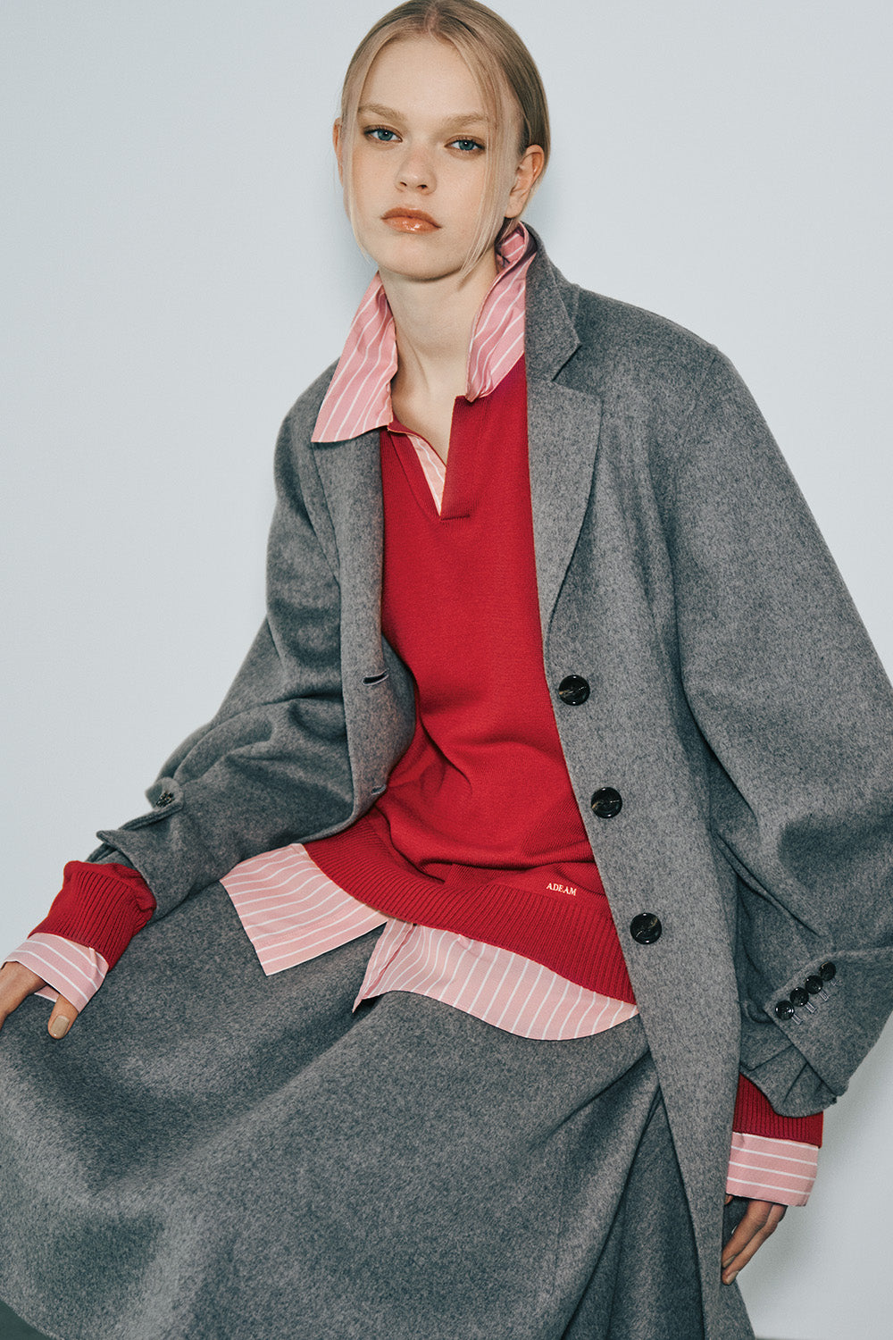 The model wears an oversized gray wool coat open over a red V-neck knit sweater and a pink-and-white striped shirt, with the shirt collar, cuffs, and hem showing, and a gray skirt.